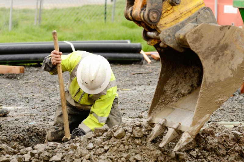 Worker in hi-vis digging alongside an excavator on a job site